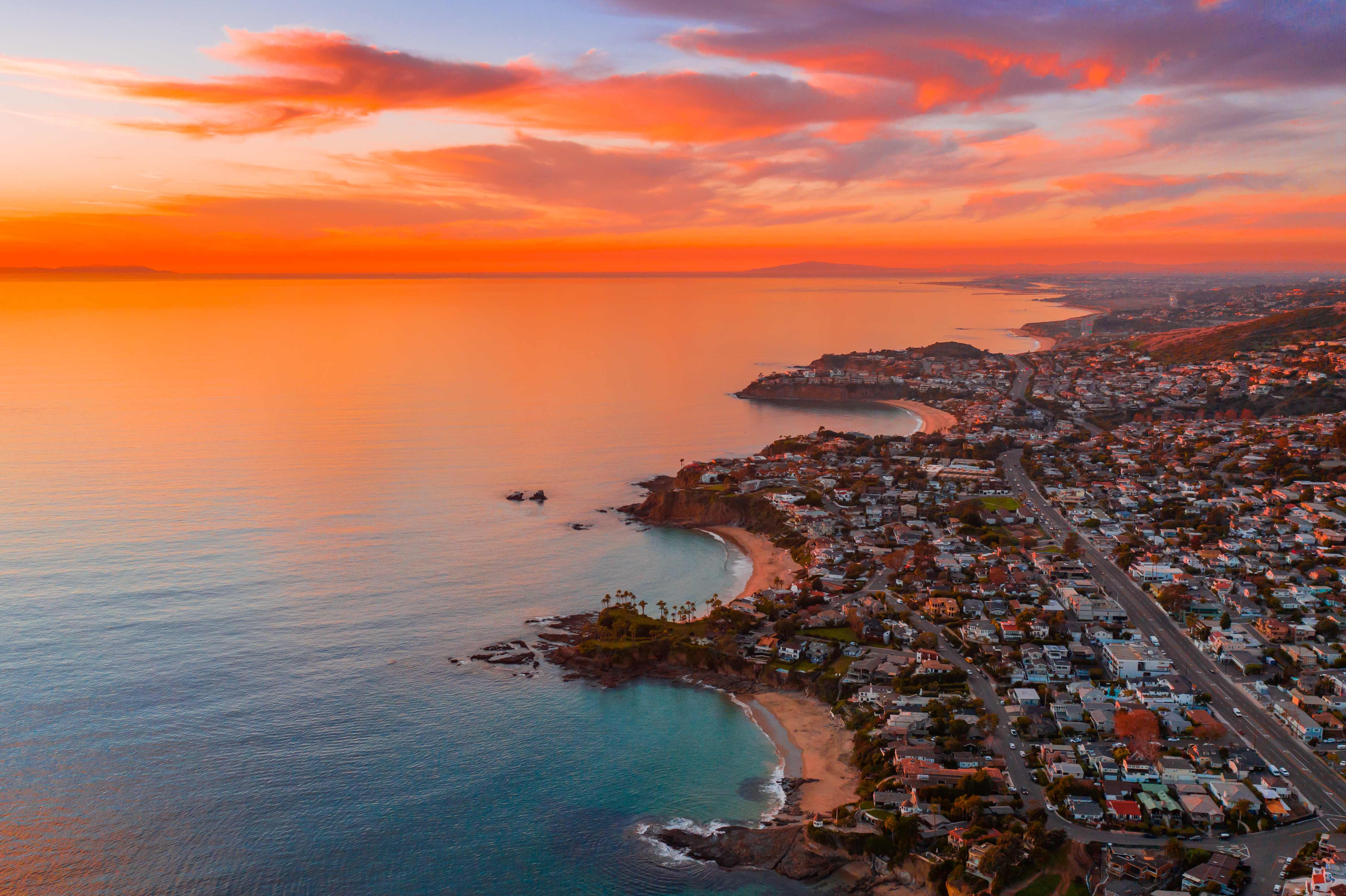 Stunning aerial sunset view of Laguna Beach coastline with vibrant orange and pink sky, turquoise ocean, and coastal homes